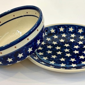 May include: Ceramic bowl and plate set with a patriotic star pattern. The bowl has a blue rim with white and brown accents, and the plate is covered in white stars on a dark blue background. The set is ideal for serving food.