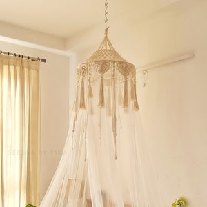 May include: A white canopy bed with a macrame canopy hanging from the ceiling. The bed has a white bedspread and a grey throw blanket. There are plants on the floor next to the bed.