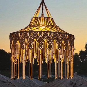 May include: A handcrafted macrame pendant light fixture. The light fixture is a warm beige color, with intricate knotting patterns and long fringe. The light shines through the macrame, creating a warm glow. The fixture is suspended from a dark brown support.