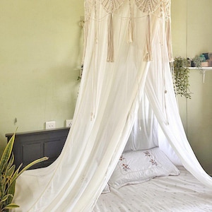 Peut inclure: Un baldaquin en macramé crème est suspendu au-dessus d'un lit avec une literie blanche. Le baldaquin a un design circulaire avec des glands suspendus. Le lit a un oreiller blanc avec un motif floral. Une plante en pot est visible dans le coin.