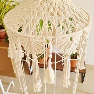 Puede incluir: Una pantalla de l&aacute;mpara de macram&eacute; color crema con intrincados patrones de nudos, una estructura escalonada con marcos circulares y largos flecos colgantes. El texto SEESEA BY FIDA es visible en la parte superior del dise&ntilde;o tejido.