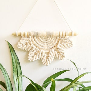 May include: A cream-colored macrame wall hanging with a half-circle design and a wooden dowel at the top. The hanging is attached to the wall with a string. A green plant with long leaves is in the foreground.