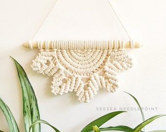 May include: A cream-colored macrame wall hanging with a half-circle design and a wooden dowel at the top. The hanging is attached to the wall with a string. A green plant with long leaves is in the foreground.