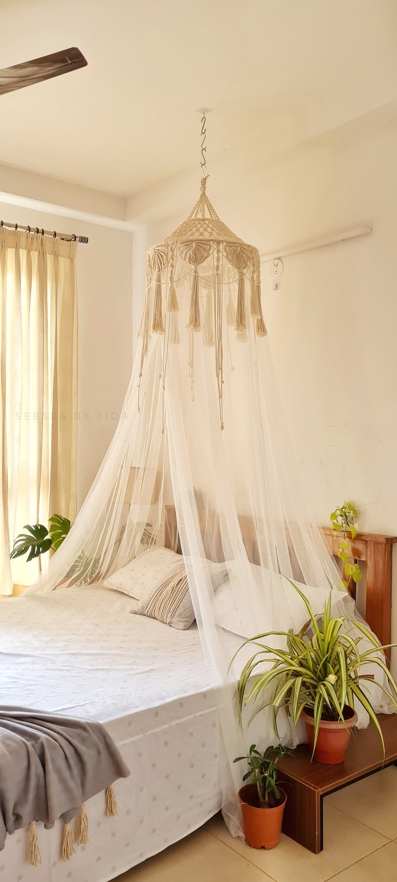 Puede incluir: Un dosel de cama de macram&eacute; color crema con una parte superior c&oacute;nica, patrones de nudos intrincados y borlas colgantes. El dosel incluye una red blanca larga y transparente que cae hacia abajo, creando un suave cerramiento sobre la cama.