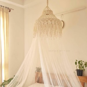 May include: A cream-coloured macrame canopy light fixture hangs above a bed, with a sheer white mosquito net draped below. The fixture has a woven design with dangling fringe. The text "SEESEA BY FIDA" is visible on the wall.