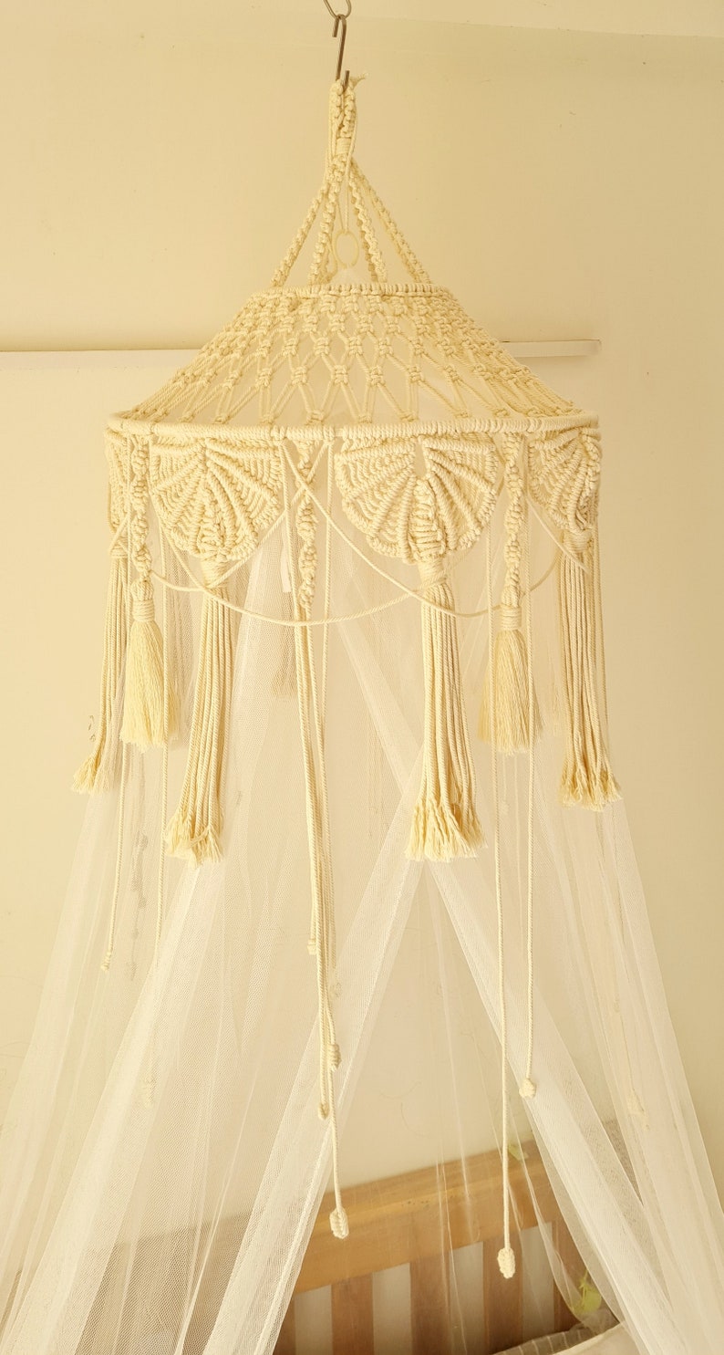 Puede incluir: Un dosel de macram&eacute; color crema con una parte superior c&oacute;nica de intrincados patrones de nudos, un marco circular adornado con motivos de macram&eacute; en forma de abanico y largas borlas colgantes. El dosel incluye una red blanca transparente.