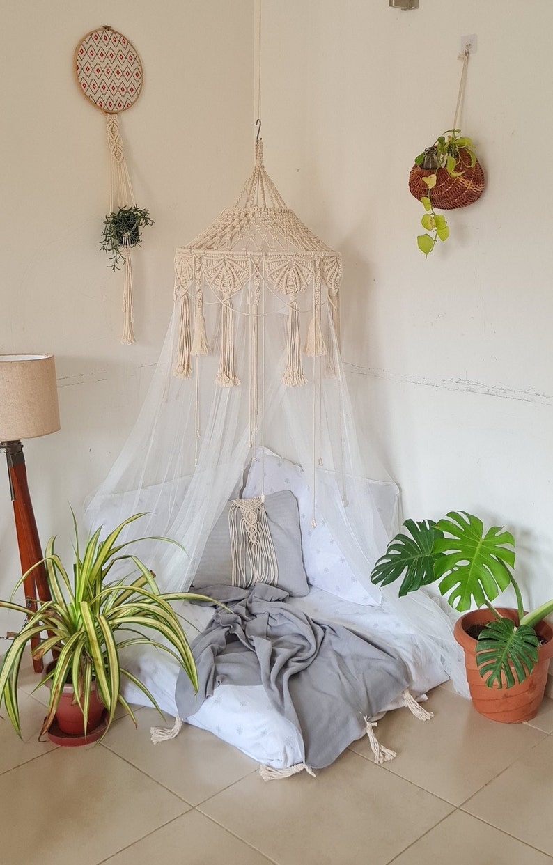 Puede incluir: Un dosel de macram&eacute; color crema con intrincados patrones de nudos y borlas colgantes, suspendido del techo. El dosel cubre un colch&oacute;n de suelo con una s&aacute;bana blanca estampada, una almohada gris y una manta gris sobre la superficie.