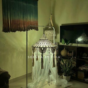 Puede incluir: Una l&aacute;mpara de dosel de macram&eacute; con parte superior c&oacute;nica, patrones de nudos intrincados y borlas colgantes, suspendida de un soporte de metal. La estructura presenta un drapeado de tela blanca transparente y el texto SEE SEA BY FIDA impreso.