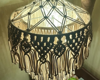 Könnte beinhalten: Beige Makramee-Pendelleuchte mit dekorativem Fransenbesatz. Die Lampe hat ein gewebtes Design und hängt an einem geflochtenen Kabel. Das Licht ist eingeschaltet und erzeugt einen warmen Schein. Die Lampe hängt in der Nähe eines Holzastes.