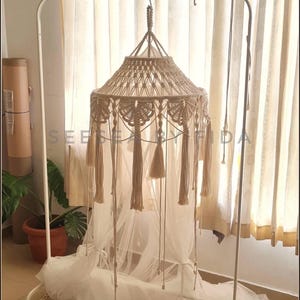 Puede incluir: Un dosel de macram&eacute; beige con intrincados patrones de nudos y borlas colgantes, suspendido de un perchero de metal blanco. El dosel incluye una tela blanca transparente que se acumula en la base, con el texto SEE BY FIDA visible.
