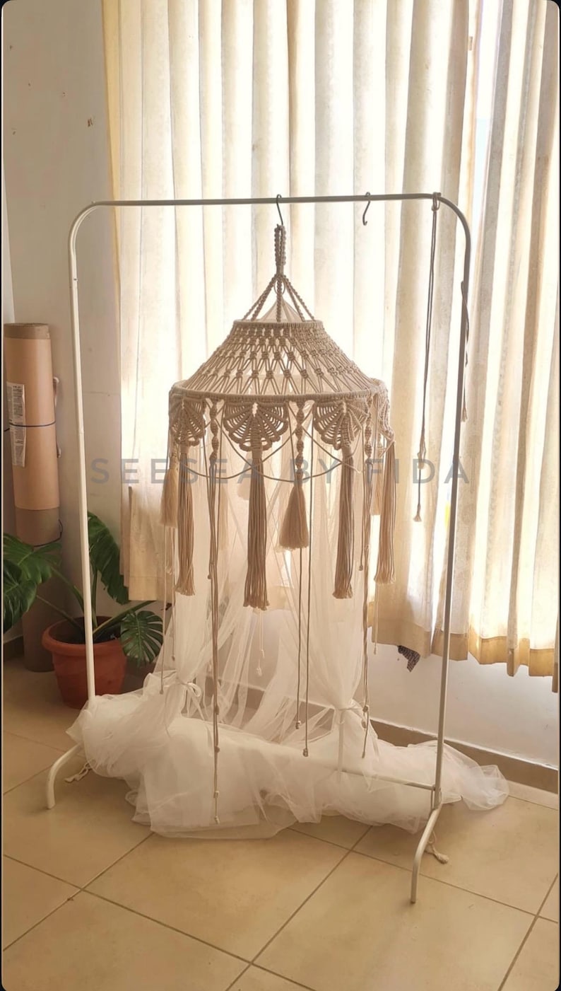 Puede incluir: Un dosel de macram&eacute; beige con intrincados patrones de nudos y borlas colgantes, suspendido de un perchero de metal blanco. El dosel incluye una tela blanca transparente que se acumula en la base, con el texto SEE BY FIDA visible.