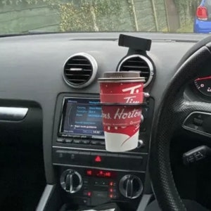 以下が含まれることがあります： 赤と白の Tim Hortons のコーヒーカップが、黒い車のカップホルダーに収まっています。 カップホルダーは車のダッシュボードに取り付けられています。 車内はグレーと黒です。