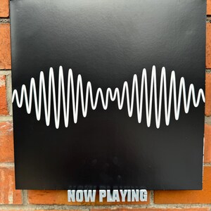 May include: A black square sign with a white waveform graphic and the text "NOW PLAYING" in silver letters.