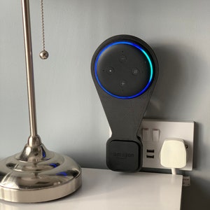 May include: A black wall mount for an Amazon Echo Dot smart speaker. The mount has a small 'amazon' logo on the bottom and a blue light ring around the speaker. The mount is plugged into a white electrical outlet.
