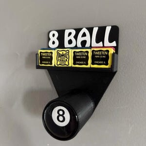 Op de afbeelding: Zwarte 8-ball houder met een plankje voor krijt. De houder heeft de woorden "8 BALL" in witte letters. Het plankje bevat vier gele krijtdozen met de tekst "TWEETEN FIBRE CO INC CHICAGO, IL" en "TRIANGLE CHALK".