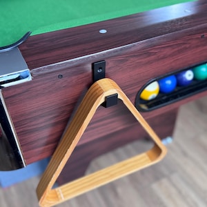 May include: A close-up of a pool table with a green felt surface and a dark wood finish. A wooden triangle rack hangs on the side, and a rack of billiard balls is visible. The table has chrome accents.