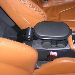 Op de afbeelding: Zwarte, gewatteerde armsteun voor in de auto, geïnstalleerd tussen twee bruinleren autostoelen. De armsteun is gemonteerd op een zwart metalen frame. De autostoelen hebben decoratieve stiksels.