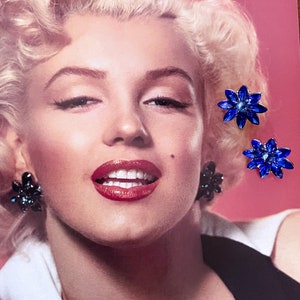 May include: A close-up of a woman's face with three pairs of blue and black flower-shaped earrings. The earrings are made of rhinestones and have a silver setting.
