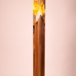 May include: A tall, wooden floor lamp with a rectangular base. The lamp has a clear resin section in the middle with gold accents. The lamp is lit and casting a warm glow.