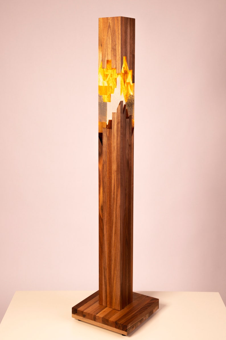 May include: A tall, wooden floor lamp with a rectangular base. The lamp has a clear resin section in the middle with gold accents. The lamp is lit and casting a warm glow.