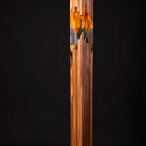May include: A tall, wooden floor lamp with a rectangular base. The lamp is made of multiple pieces of wood that are stacked and glued together. The lamp has a clear glass cylinder in the middle, which is filled with a golden resin. The resin has a unique, abstract pattern.