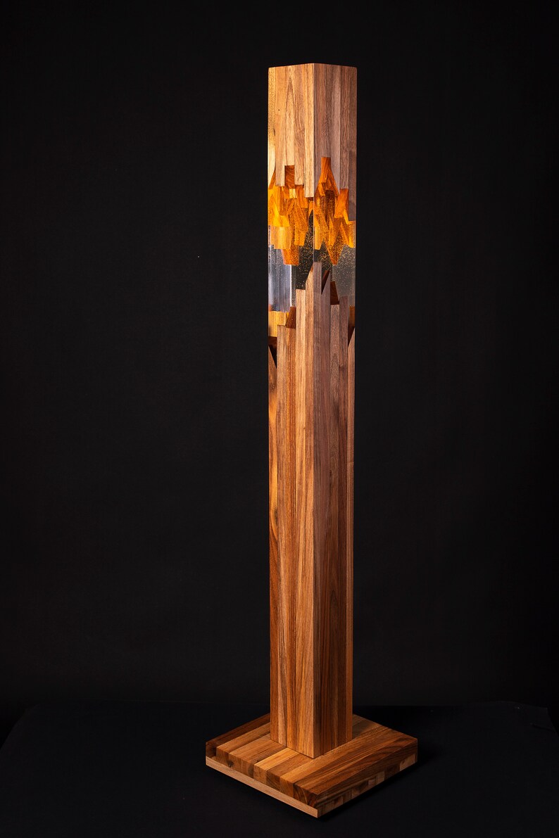 May include: A tall, wooden floor lamp with a rectangular base. The lamp is made of multiple pieces of wood that are stacked and glued together. The lamp has a clear glass cylinder in the middle, which is filled with a golden resin. The resin has a unique, abstract pattern.