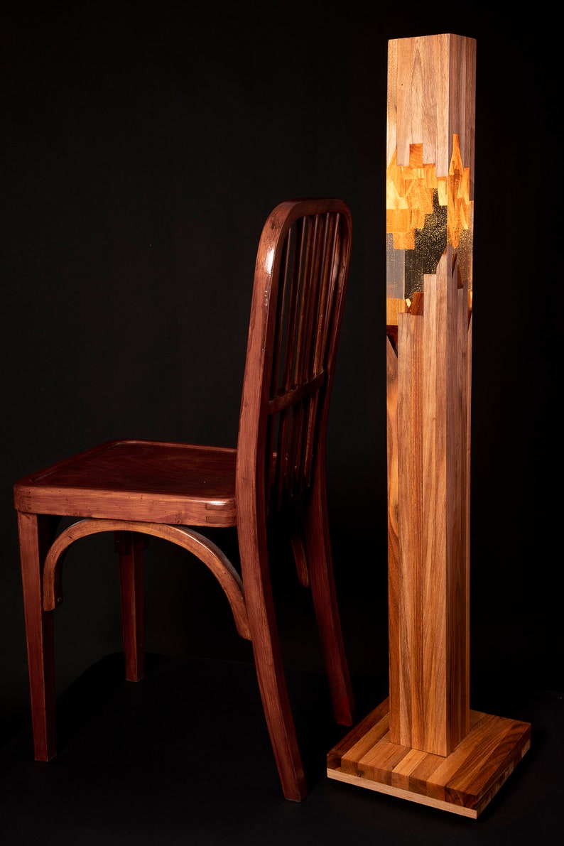 May include: A wooden chair with a dark brown finish sits next to a tall, wooden floor lamp. The lamp has a clear glass section in the middle with gold leaf accents.