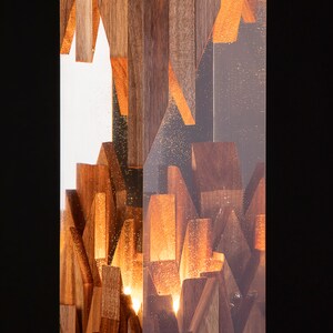 May include: A rectangular wooden lamp with a clear resin exterior. The wood is arranged in a geometric pattern and illuminated from within, creating a warm glow.
