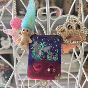 May include: A whimsical necklace featuring a purple and red game console pendant with a heart, glitter, and a clear top. The necklace also includes a troll doll, a crown, and a pink fairy. The chain is gold-colored.