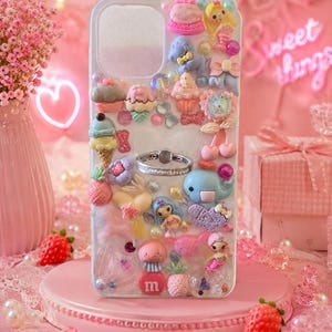 Könnte beinhalten: Eine weiße Smartphone-Hülle, verziert mit einer Vielzahl bunter Miniatur-Anhängern wie Cupcakes, Eiswaffeln, einem blauen Wal, einer Meerjungfrau, einer Qualle, einem roten Buchstaben m und einem silbernen Ringhalter in der Mitte.