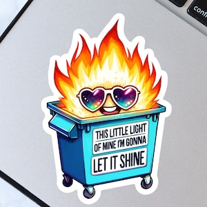 Puede incluir: Un contenedor de basura azul con llamas en la parte superior y una cara sonriente con gafas de sol. El contenedor tiene una pegatina blanca que dice "This little light of mine I'm gonna let it shine."