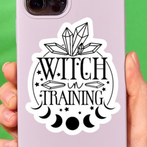 May include: A purple phone case with a black and white sticker that says "Witch in Training" with a crescent moon and stars around it.