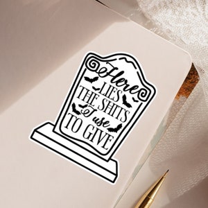 May include: Black and white sticker of a tombstone with the text "Here Lies The Shits I Use To Give".