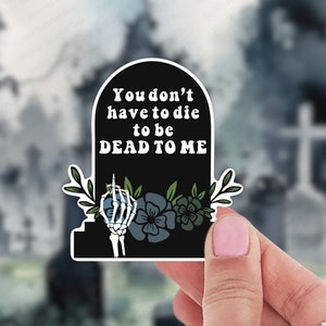 May include: Black and white sticker of a tombstone with the text "You don't have to die to be dead to me" and a skeleton hand giving the middle finger. The sticker is surrounded by green leaves and flowers.