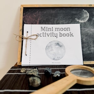 May include: A white "Mini moon activity book" with a watercolor moon illustration, tied with twine, rests against a chalkboard. A wooden magnifying glass and small rocks are in the foreground.