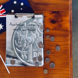 May include: A clipboard with the title "Australian currency math workbook" and a close-up image of an Australian one dollar coin.  There are several Australian coins scattered on a wooden surface.
