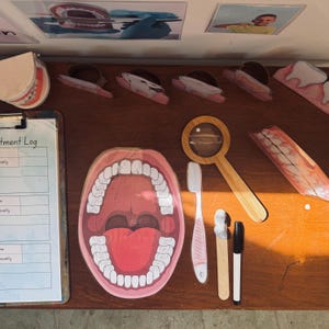 Puede incluir: Una mesa de madera con una variedad de herramientas de salud dental y ayudas educativas. Hay una boca grande abierta con dientes, una lupa, un cepillo de dientes, un tubo de pasta de dientes, un palillo de dientes, un marcador negro y un portapapeles con el encabezado "Registro de tratamiento".