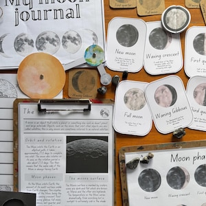 May include: A collection of educational materials about the moon, including a journal, phase cards, and informational sheets. The cards illustrate the moon phases, and the journal has a moon phase chart. The items are arranged on a wooden surface.