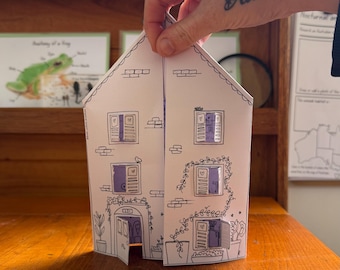 Op de afbeelding: Een wit papieren huisje met zwarte lijntekeningen van ramen, deuren en klimplanten. De structuur heeft een puntdak en bevat verschillende uitgesneden delen die openen om paarse details aan de binnenkant van het papier te onthullen.