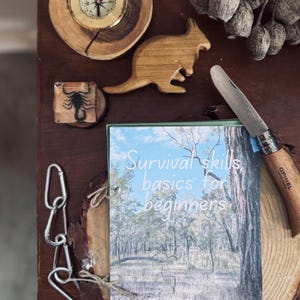 以下が含まれることがあります： 暗い木製の表面に、サバイバルをテーマにしたアイテムのコレクション。コンパス、木製のカンガルーの置物、サソリの樹脂ペーパーウェイト、ナイフ、そして「Survival skills basics for beginners」というタイトルの本が含まれています。