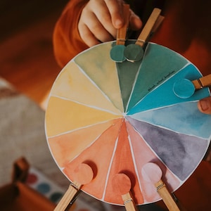 Peut inclure: Un nuancier circulaire composé de douze segments aquarellés distincts dans des tons de jaune, orange, rouge, violet, bleu et vert. Le disque porte l'inscription @bush_school_kreatives et est maintenu par plusieurs pinces à linge en bois.