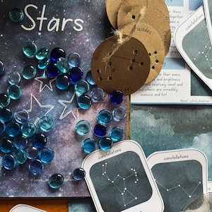 Puede incluir: Una vista cenital presenta un tema celestial con la palabra "Stars" en blanco. Cuentas de vidrio azules y transparentes se dispersan alrededor de contornos de estrellas. Etiquetas redondas marrones con diseños de constelaciones cuelgan de hilo. Tarjetas "constellations".