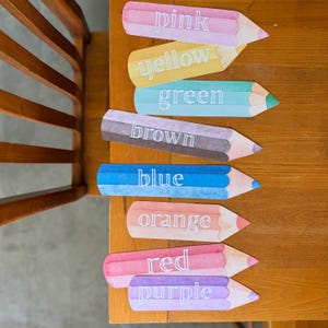 Op de afbeelding: Een verzameling kleurrijke potloodvormige uitsparingen, elk met een andere kleurnaam: roze, geel, groen, bruin, blauw, oranje, rood en paars. De uitsparingen zijn gerangschikt op een houten oppervlak.