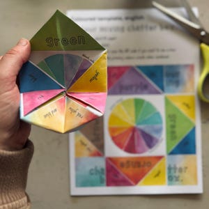 May include: A paper fortune teller toy featuring various colors and text labels including green, blue, yellow, red, and blue, and red and yellow, held above a printed template sheet with a color wheel and additional text labels.