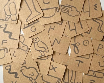 Puede incluir: Un conjunto de 26 cartas con fondos de papel marrón y dibujos de tinta negra de letras y símbolos. Las cartas están dispuestas en un patrón aleatorio.