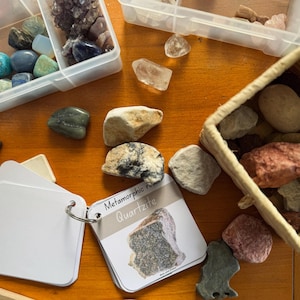 Peut inclure: Une collection de roches et de minéraux, dont du quartz, exposée sur une surface en bois. L'image comprend de petits contenants en plastique et un panier en osier rempli de pierres. Une carte avec le texte "Metamorphic rocks Quartzite" est également présente.