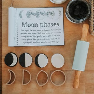 Pode incluir: Um conjunto de cortadores de argila com fases lunares, discos de argila circulares, um rolo de massa azul claro e um cartão educativo intitulado Moon phases. O cartão explica as 8 fases lunares e o ciclo de 29,5 dias. Os cortadores são bege.