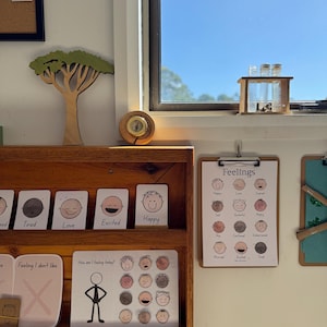 Puede incluir: Estantería de madera con tarjetas de emociones: "Cansado", "Amor", "Emocionado" y "Feliz". Un portapapeles muestra una tabla de "Sentimientos". Decoración de árbol de madera y un pequeño reloj.
