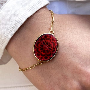 Może przedstawiać: Złota bransoletka z łańcuszkiem z czerwonym owalnym wisiorkiem z czarnym wzorem kwiatowym.