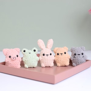 Op de afbeelding: Een verzameling van vijf kleine, gehaakte dierenfiguren in pastelkleuren, waaronder roze, groen, perzik en grijs. De figuren, een varken, een kikker, een konijn, een beer en een kat, staan op een roze dienblad.